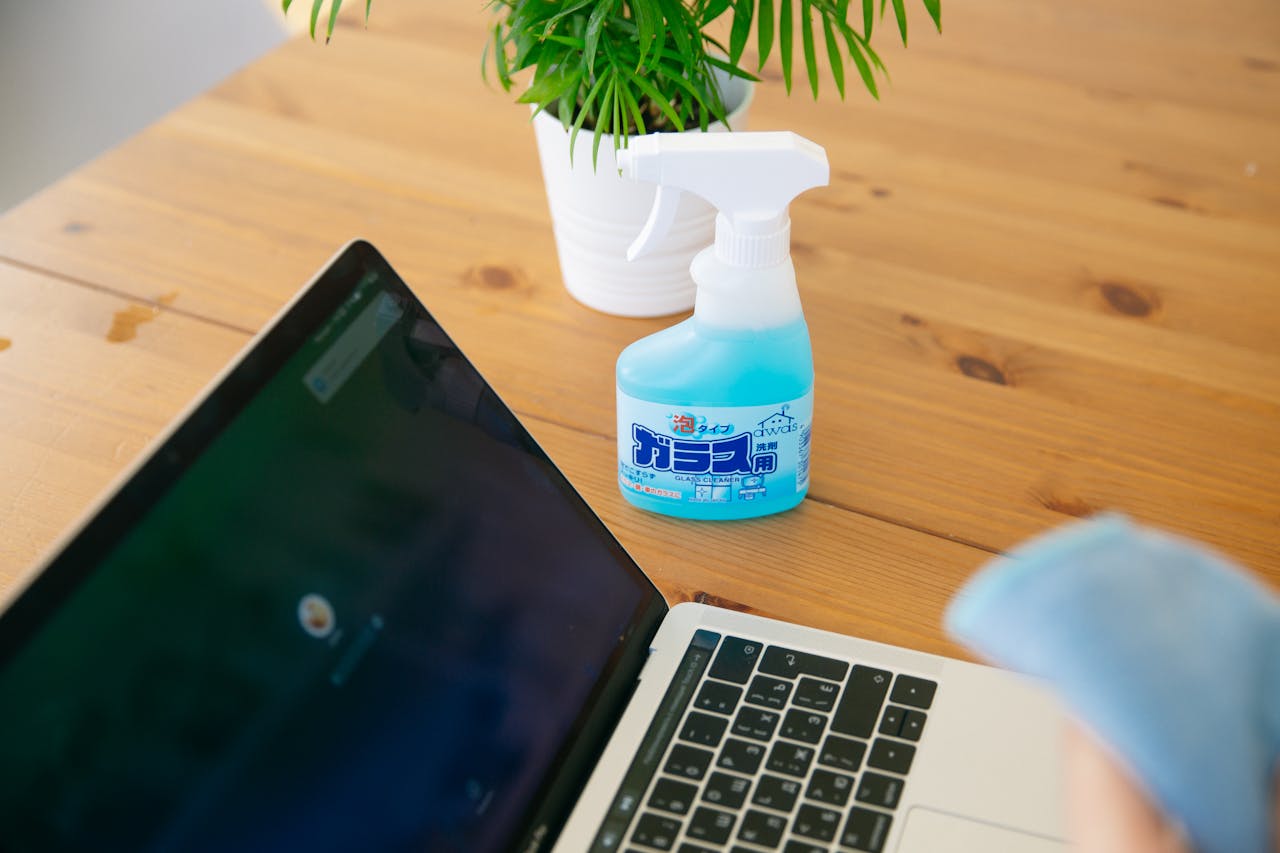 A laptop with a spray bottle and plant on a wooden desk, perfect for remote work and cleaning themes.